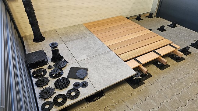 Outdoor terrace construction display featuring wooden decking planks, stone tiles, and plastic paving pedestals