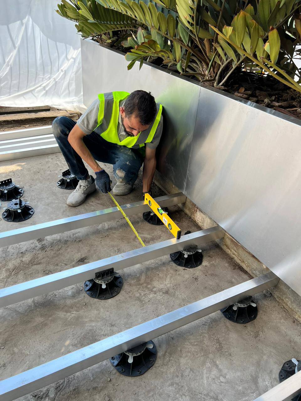 Worker using a tape measure and spirit level to align aluminum joist framing on raised deck pedestals.