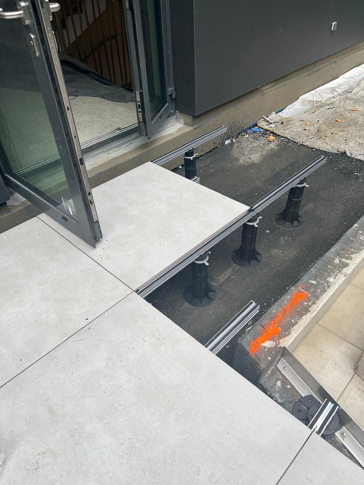 Montaż przy drzwiach tarasowych z płytkami porcelanowymi na zewnątrz ułożonymi na legarach aluminiowych i regulowanych plastikowych wspornikach tarasowych.