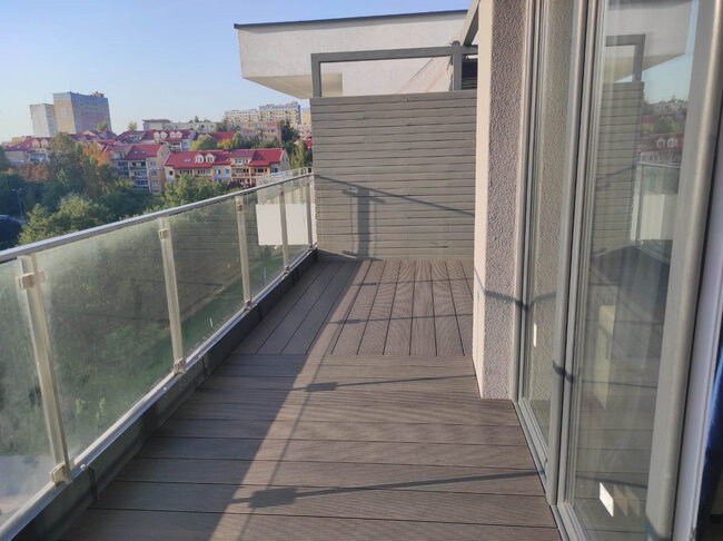 Nowoczesny balkon apartamentowy z deskami kompozytowymi, szklanymi balustradami i przegrodą zapewniającą prywatność.