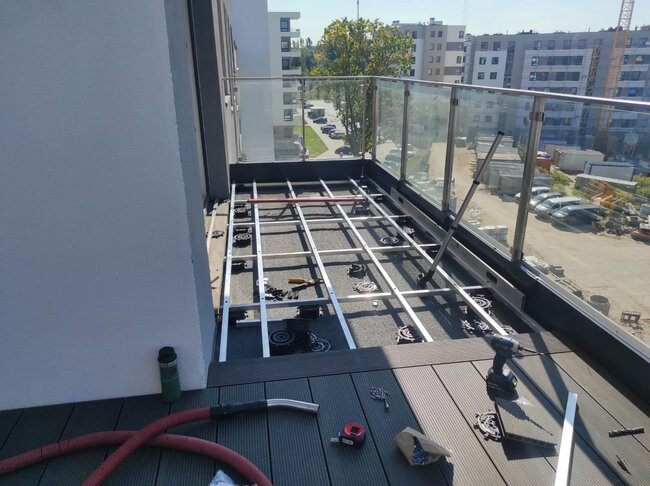 Montaż tarasu balkonowego w trakcie realizacji z widoczną konstrukcją legarów aluminiowych, wspornikami regulowanymi i deskami kompozytowymi.