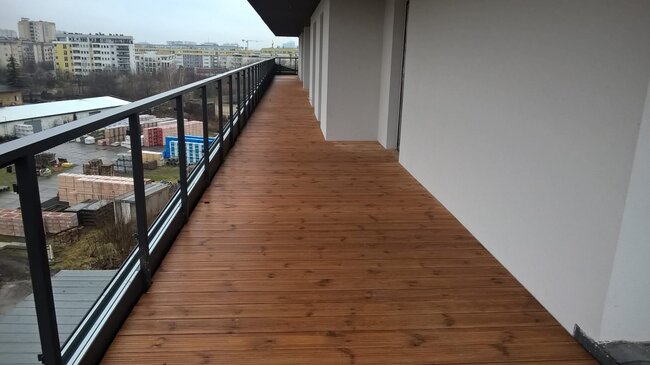 Podniesiony taras drewniany na długim balkonie apartamentu ze szklanymi i stalowymi balustradami.