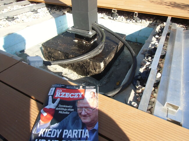 Kabel elektryczny poprowadzony wzdłuż podstawy słupka i leżącej poniżej struktury żwirowej pod podniesionym tarasem wentylowanym.